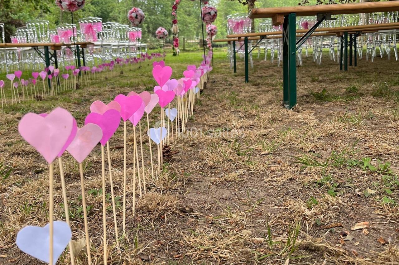 Allée décorée de cœurs en papier rose et violet menant à une cérémonie en plein air avec des chaises et des arches fleuries.