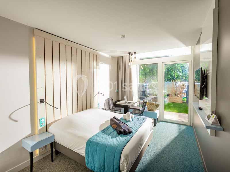 Chambre lumineuse avec lit double, bureau, télévision et accès à une terrasse donnant sur un jardin verdoyant.