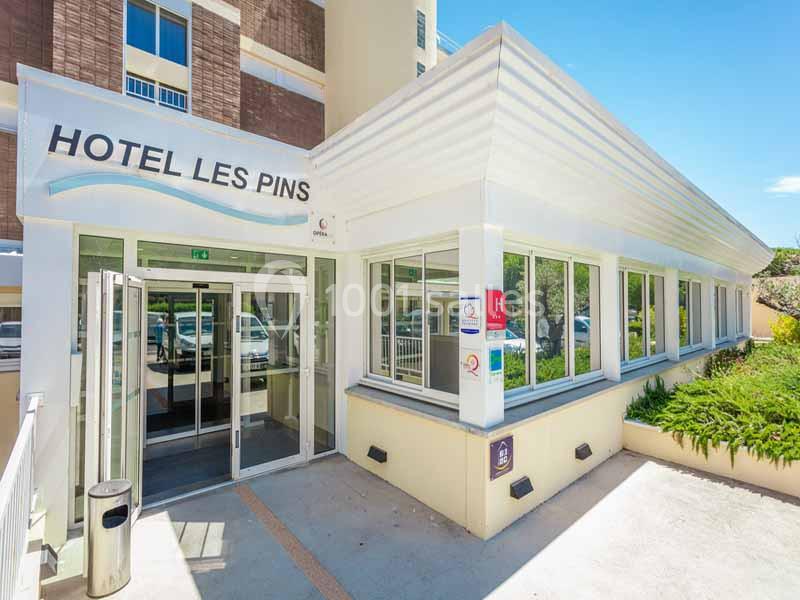 Entrée d'un hôtel avec une façade vitrée, un panneau ’Hôtel Les Pins’ et un environnement extérieur ensoleillé.