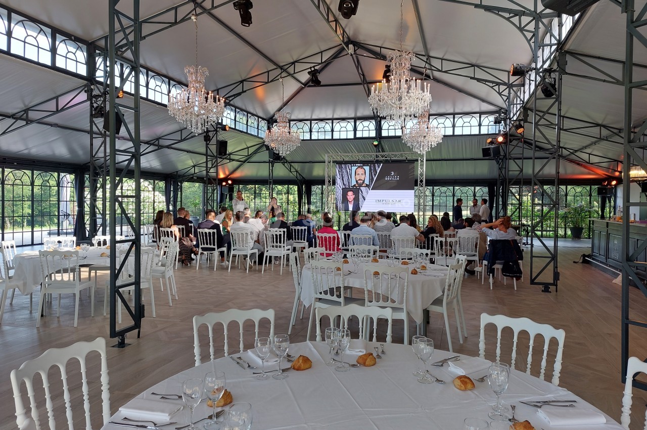 Salle de réception lumineuse avec tables dressées, invités assis et écran projetant une présentation.