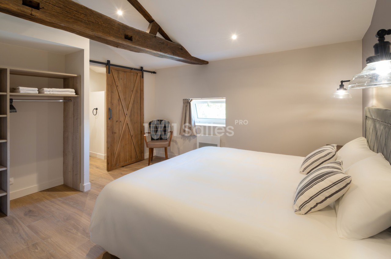 Chambre lumineuse avec lit double, poutres apparentes, porte coulissante en bois et espace de rangement ouvert.