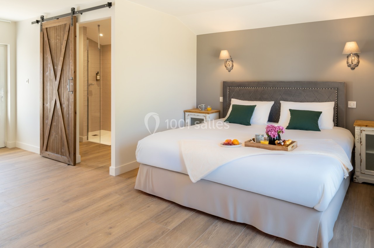 Chambre lumineuse avec un grand lit, tête de lit capitonnée, porte coulissante en bois et salle de bain attenante.
