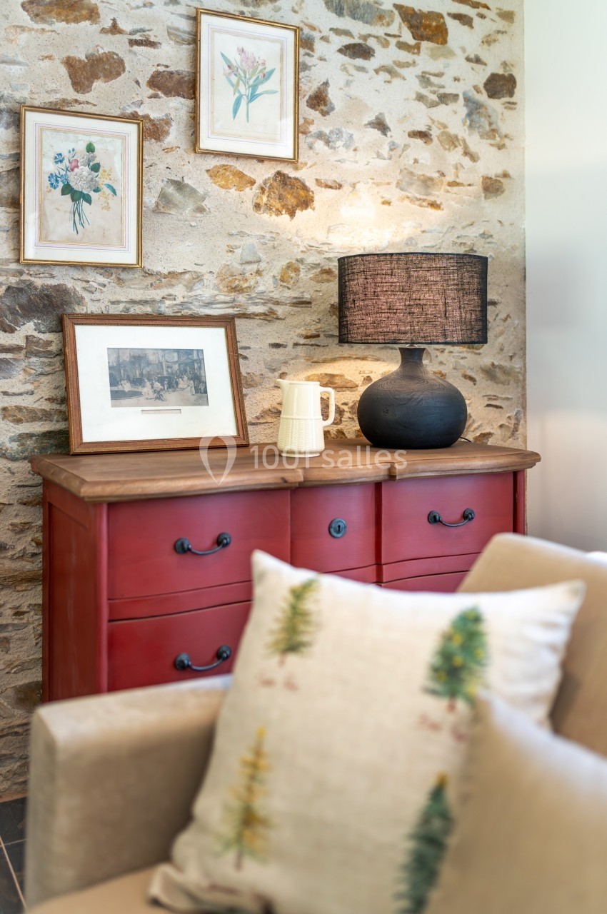 Commode rouge surmontée de cadres, lampe noire et carafe, avec un mur en pierre en arrière-plan.
