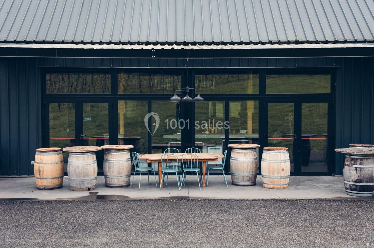 Façade d'un bâtiment avec une table en bois, des chaises métalliques et des tonneaux disposés sur une terrasse en béton.