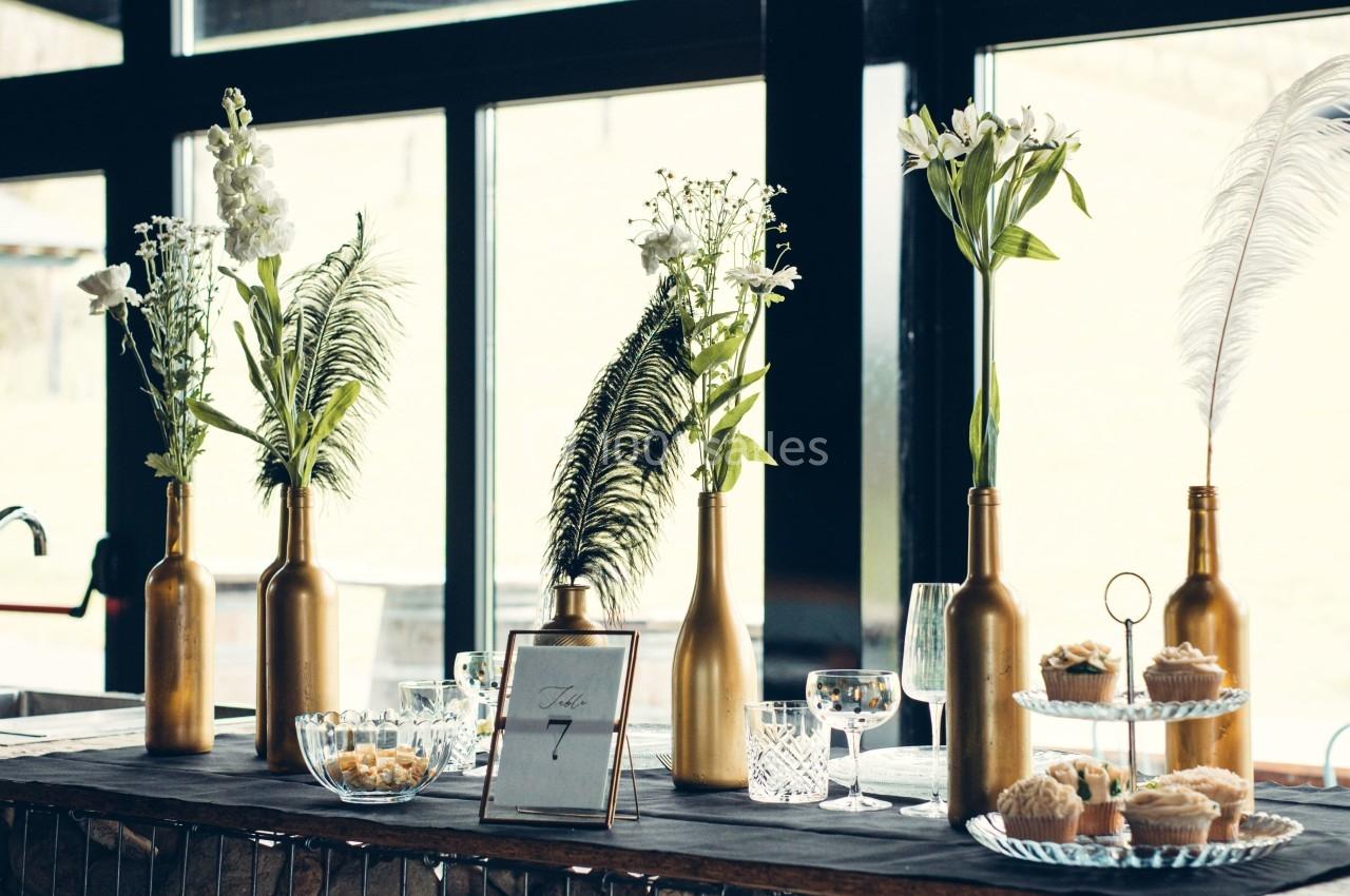 Décoration de table avec bouteilles dorées contenant des fleurs, plumes et vaisselle dans un cadre lumineux.