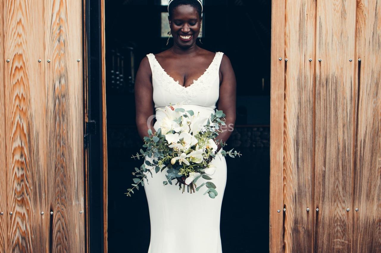 Femme en robe de mariée blanche tenant un bouquet de fleurs, debout dans l'encadrement d'une porte en bois.