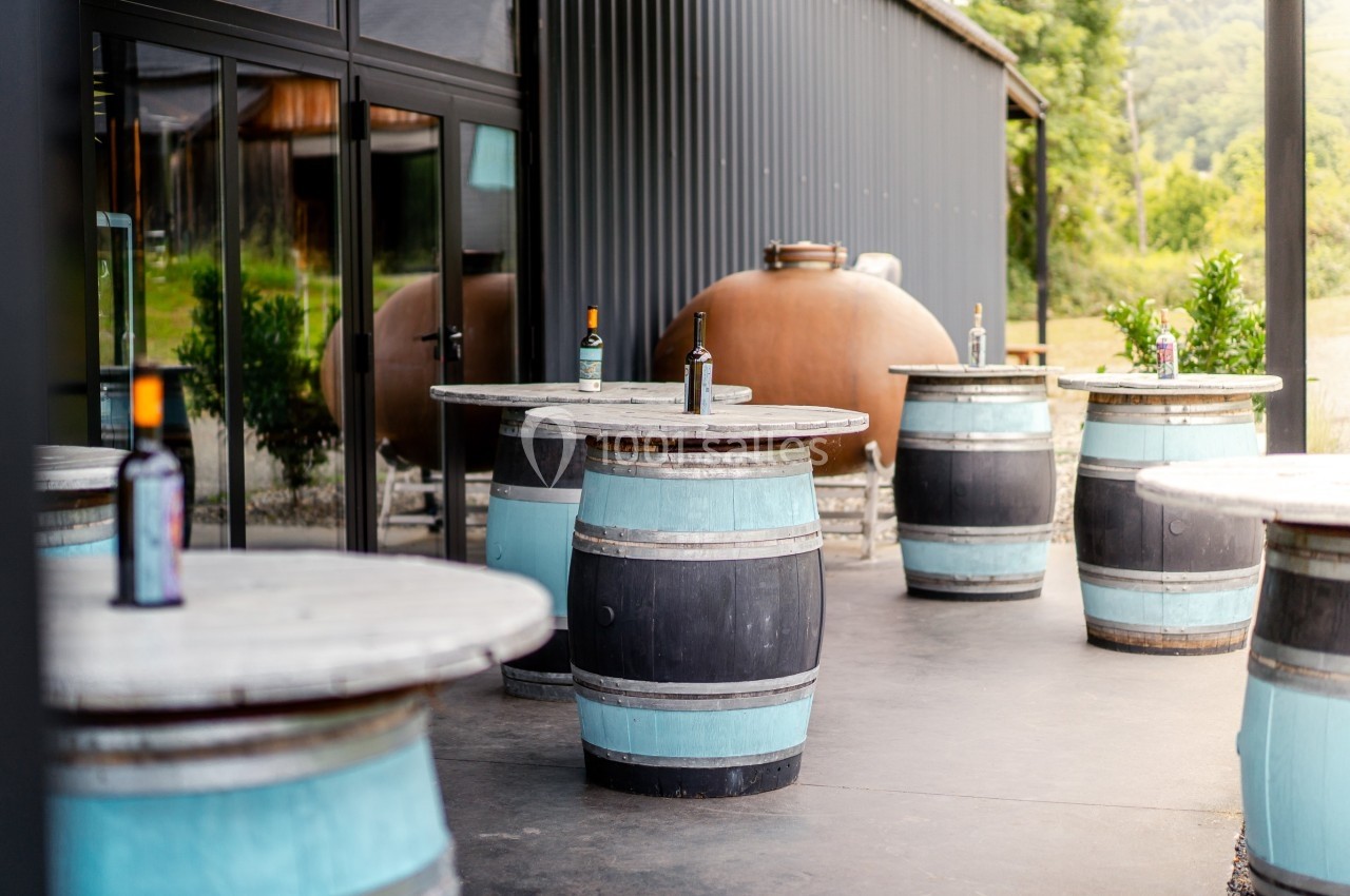 Tables en tonneaux disposées sur une terrasse extérieure près d'un bâtiment moderne, avec des bouteilles posées dessus.