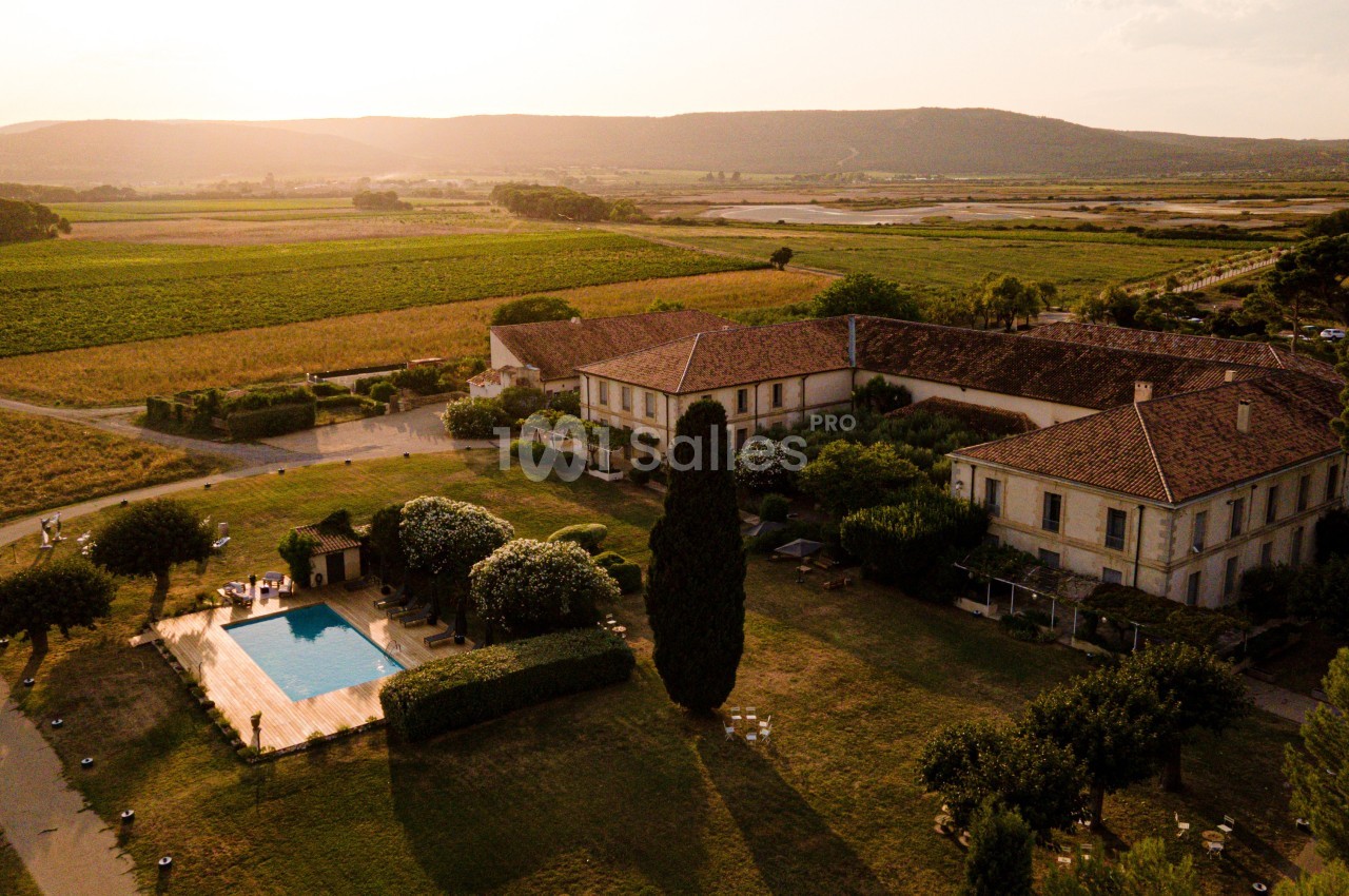 Vue aérienne d'un domaine rural avec bâtiments anciens, piscine, arbres et champs sous une lumière de coucher de soleil.