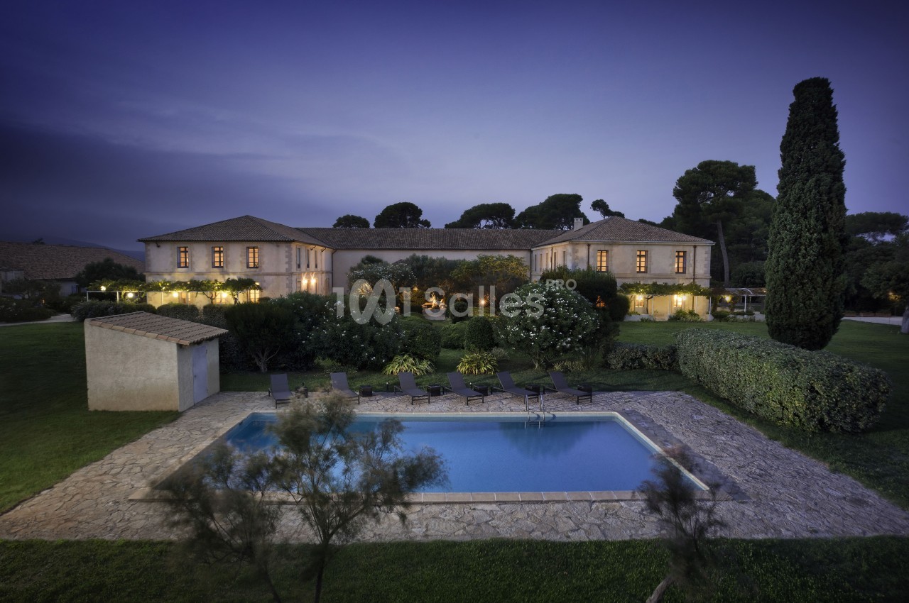 Vue d'une grande maison en pierre entourée de jardins, avec une piscine éclairée au premier plan, au crépuscule.