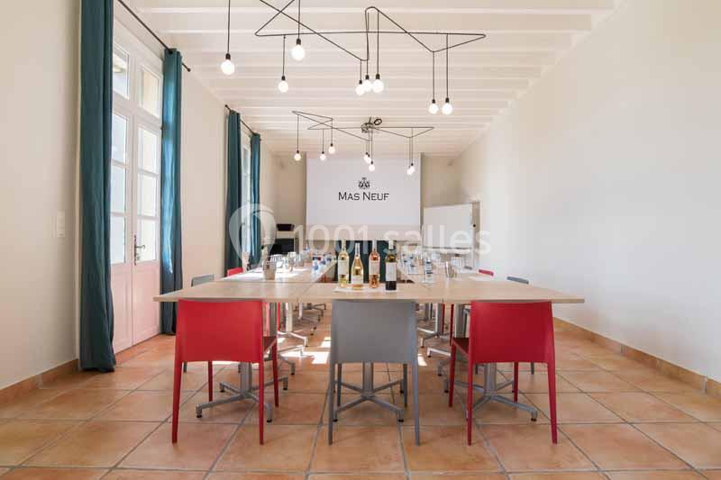 Salle de réunion lumineuse avec table rectangulaire, chaises colorées, bouteilles disposées et éclairage suspendu moderne.