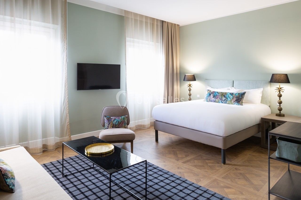 Chambre lumineuse avec lit double, fauteuil, table basse en verre, télévision murale et grandes fenêtres avec rideaux.