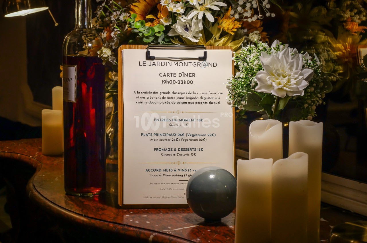 Menu de restaurant affiché sur un chevalet en bois, entouré de bougies, fleurs et bouteilles sur une table.