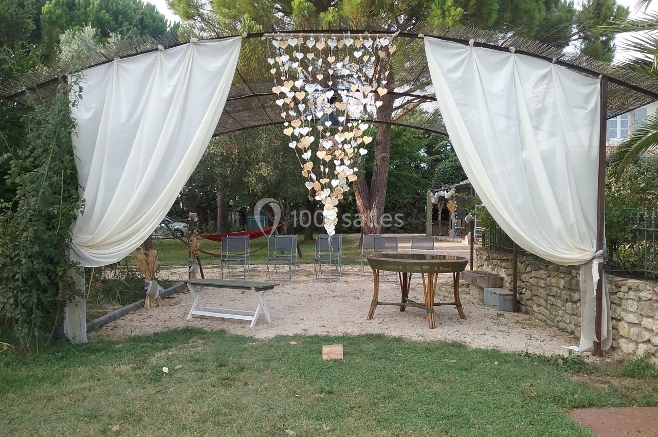 Location salle Caderousse (Vaucluse) - Domaine de la Lusignane #21 Structure en plein air avec rideaux blancs, décorations suspendues et mobilier de jardin sur sol en gravier.