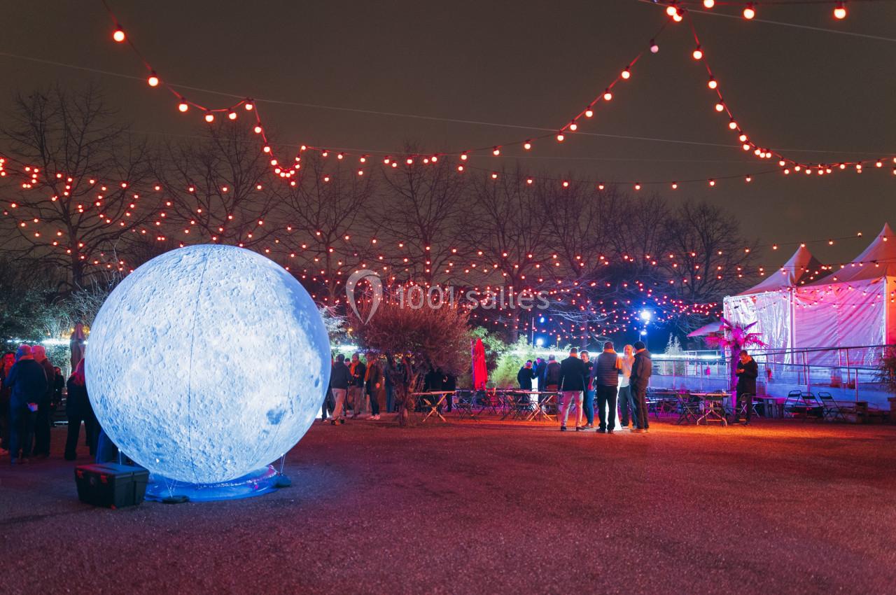 Installation lumineuse représentant une lune géante entourée de guirlandes rouges dans un espace extérieur nocturne.