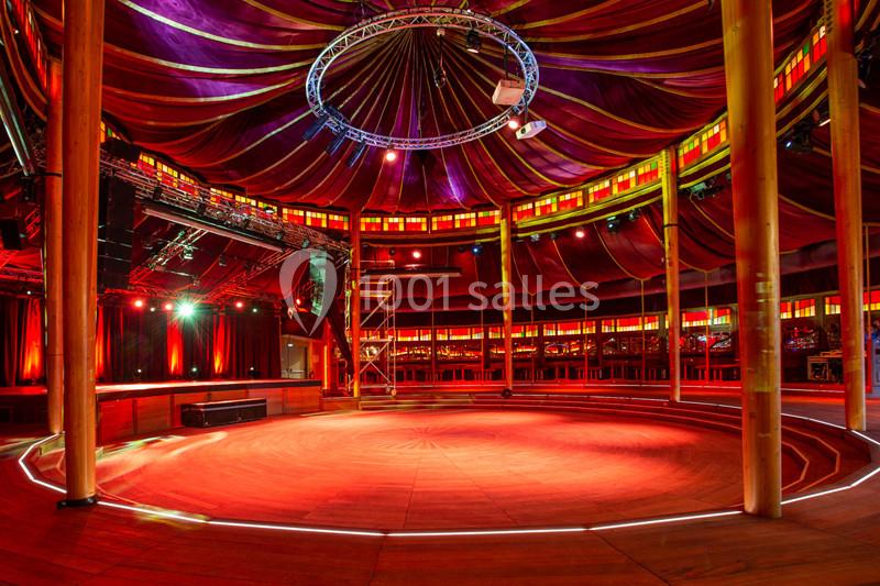Intérieur d'un chapiteau circulaire avec une scène éclairée en rouge et des gradins entourant l'espace central.
