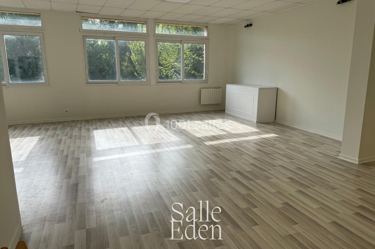 Salle vide avec parquet clair, murs blancs, grandes fenêtres laissant entrer la lumière naturelle et vue sur des arbres.