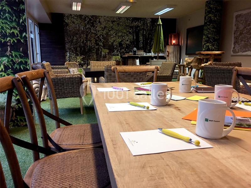 Location salle Louveciennes (Yvelines) - Campus BNP Paribas Louveciennes #14 Salle de réunion avec table en bois, chaises en osier, mugs, carnets et décor mural représentant une forêt.