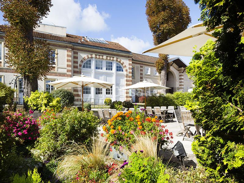 Location salle Louveciennes (Yvelines) - Campus BNP Paribas Louveciennes #8 Jardin fleuri avec terrasse aménagée, tables et parasols devant une bâtisse en briques et pierres sous un ciel bleu.