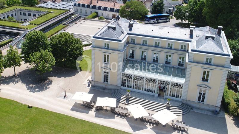 Vue aérienne d'un bâtiment historique avec terrasse aménagée, entouré de jardins et d'un parking avec un bus.