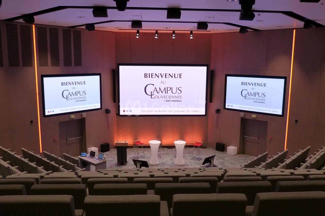Salle de conférence moderne avec sièges en gradins, écrans affichant ’Bienvenue au Campus Louvainensis’.