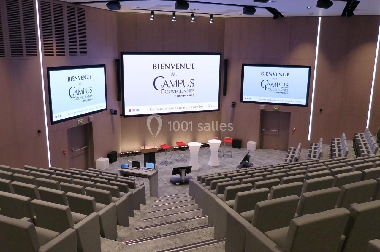 Salle de conférence moderne avec sièges gris, écrans géants affichant ’Bienvenue au Campus Louvain’.