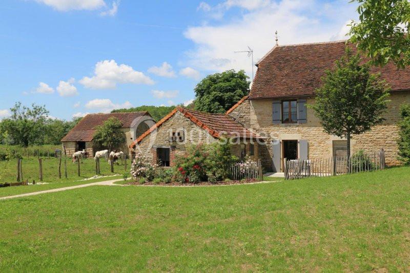 Maison en pierre avec toit en tuiles entourée de verdure, jardin fleuri et dépendances sous un ciel bleu.