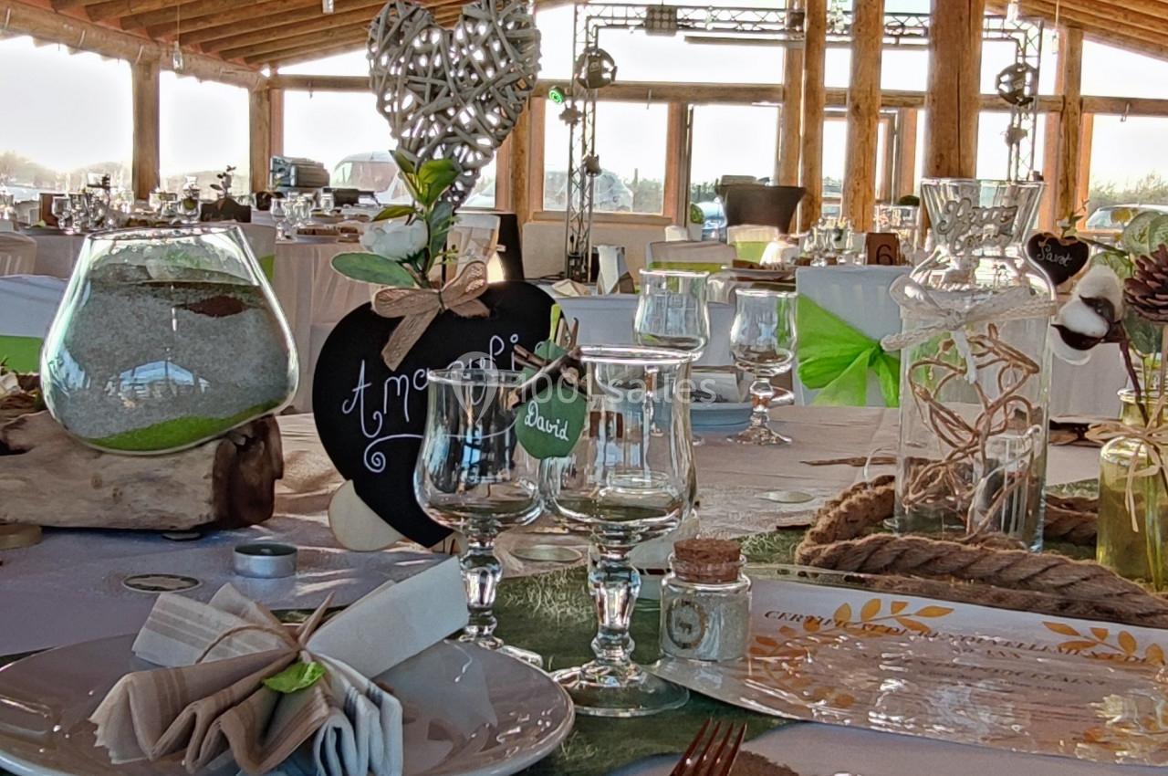 Table décorée pour un événement avec nappes blanches, couverts dorés, verres, et ornements sous un toit en bois.