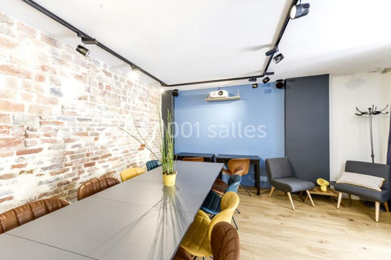 Location salle Paris 11 (Paris) - Les Rendez-vous #4 Salle lumineuse avec mur en briques, table longue entourée de chaises colorées, fauteuils et projecteur au mur.