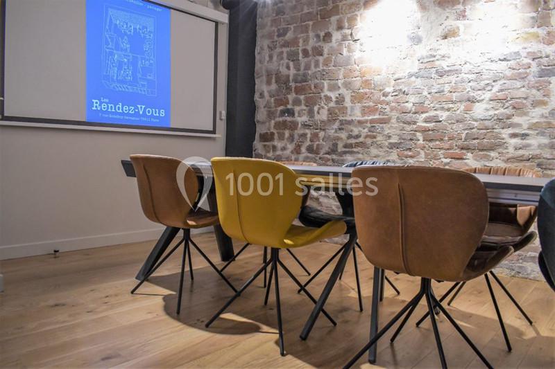 Location salle Paris 11 (Paris) - Les Rendez-vous #8 Salle de réunion avec table en bois, chaises modernes et mur en briques, écran de projection allumé.