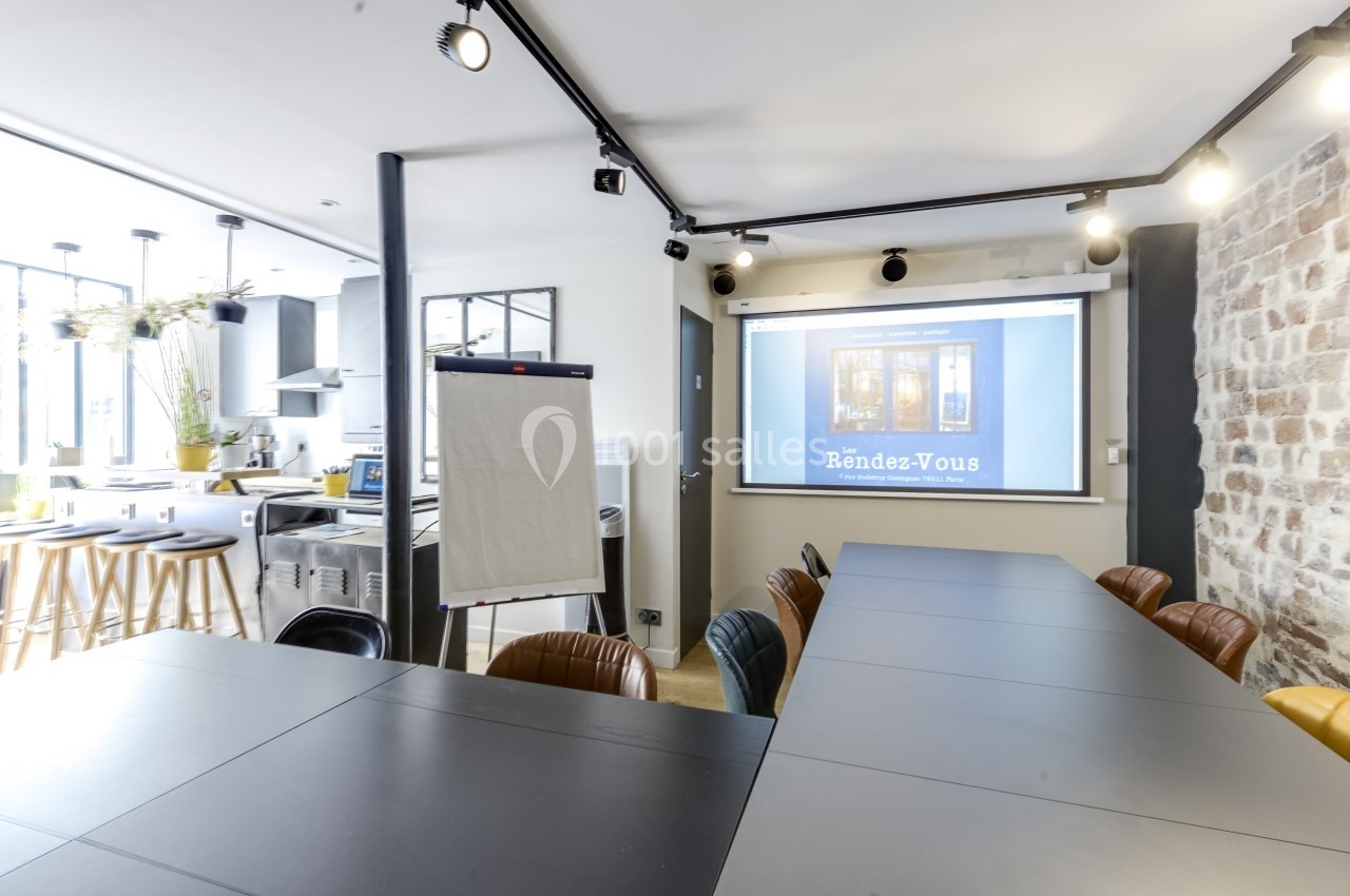 Location salle Paris 11 (Paris) - Les Rendez-vous #12 Salle de réunion moderne avec table noire, chaises, écran de projection, paperboard et cuisine ouverte en arrière-plan.