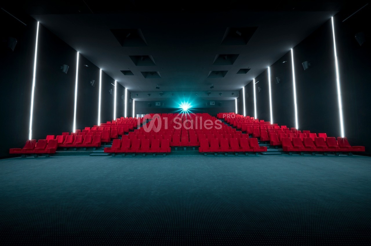 Salle de cinéma vide avec des rangées de sièges rouges et un projecteur allumé au fond.