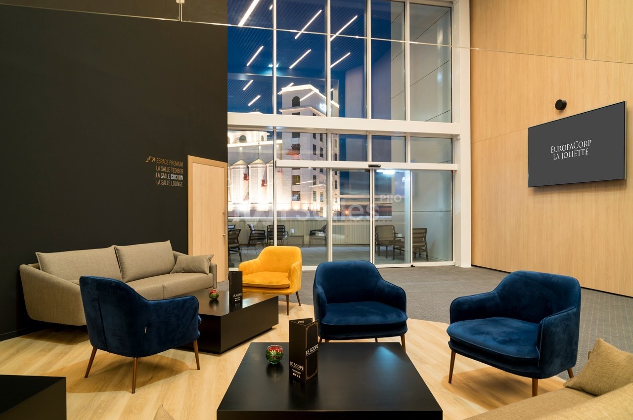 Espace lounge moderne avec fauteuils colorés, canapés et vue sur des bâtiments éclairés à travers une grande baie vitrée.