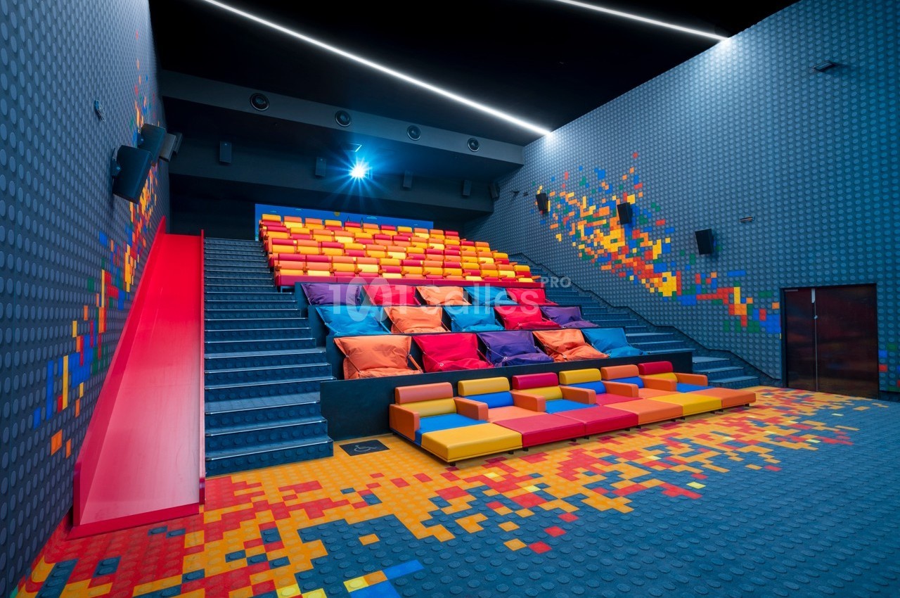 Salle de cinéma colorée avec gradins en coussins et sièges modulables, décorée de motifs pixelisés.
