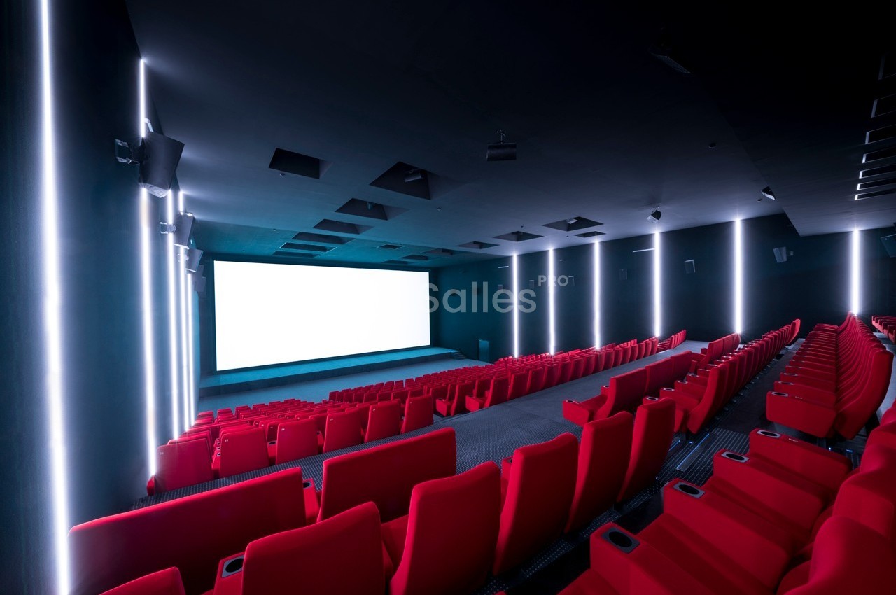 Salle de cinéma vide avec des rangées de sièges rouges et un grand écran blanc allumé.