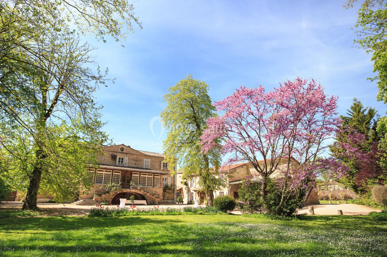 Manoir en pierre entouré d'arbres en fleurs et d'une pelouse verdoyante sous un ciel bleu clair.