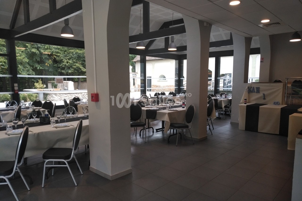 Salle de restaurant lumineuse avec tables dressées, chaises noires et grandes baies vitrées donnant sur un espace extérieur…