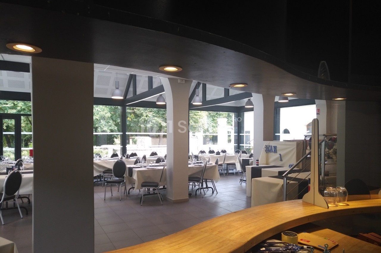 Salle de restaurant lumineuse avec tables dressées, grandes baies vitrées et vue sur un espace arboré.