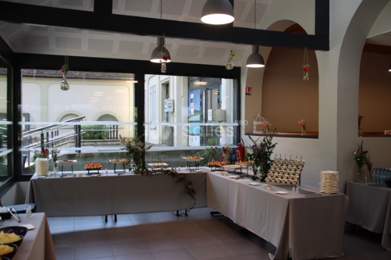 Salle lumineuse aménagée pour un buffet, avec tables dressées, décorations florales et grandes baies vitrées.