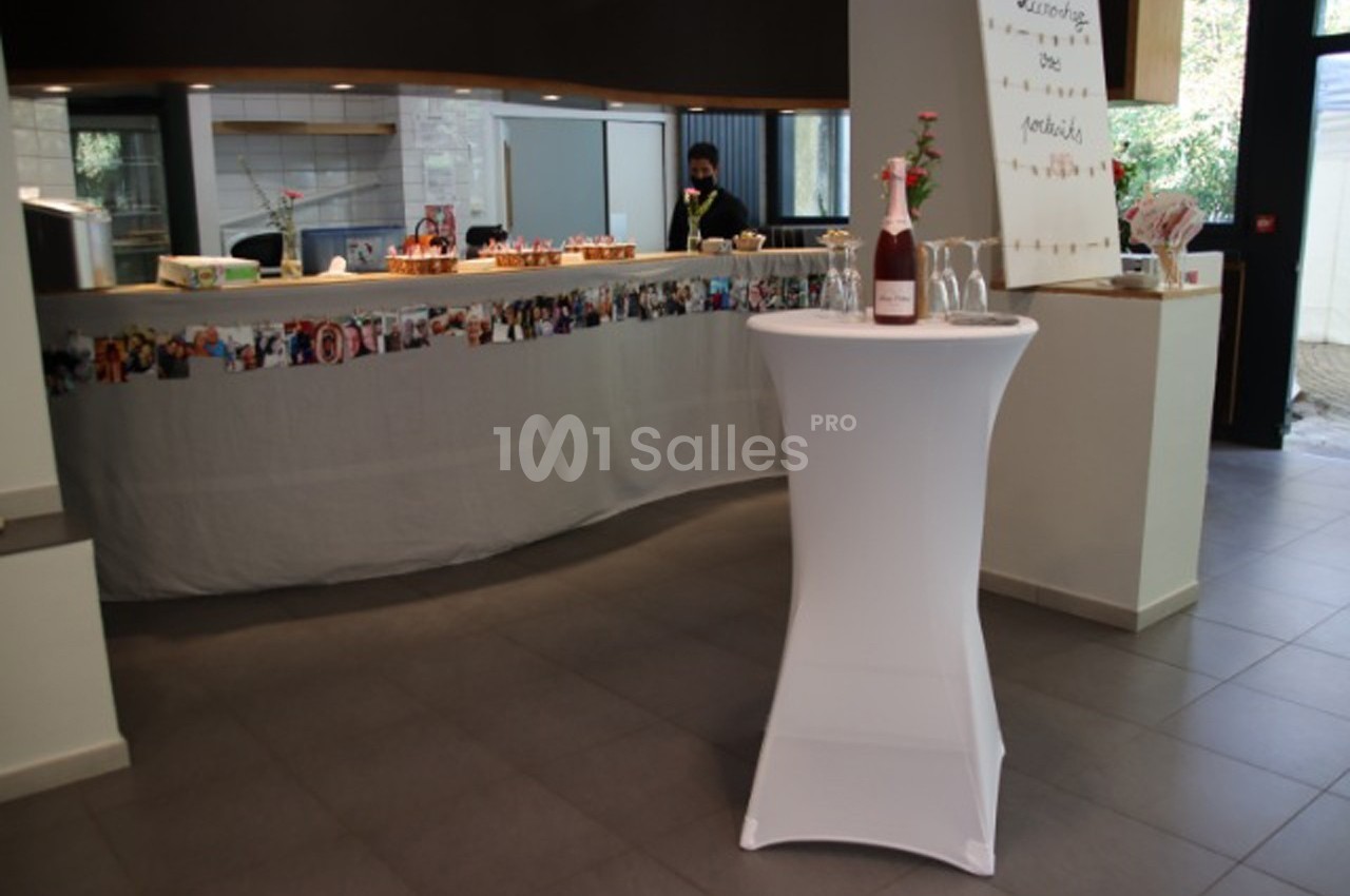 Table haute avec nappe blanche et bouteille posée dessus, devant un comptoir décoré de photos dans une salle lumineuse.