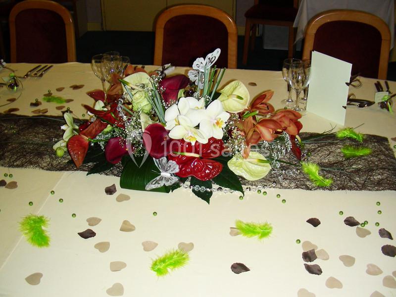 Centre de table floral avec des fleurs colorées, des plumes vertes et des décorations sur une nappe blanche.