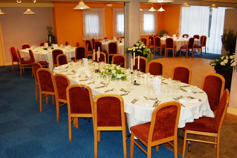 Salle de réception avec tables dressées, nappes blanches, chaises rouges et décorations florales.