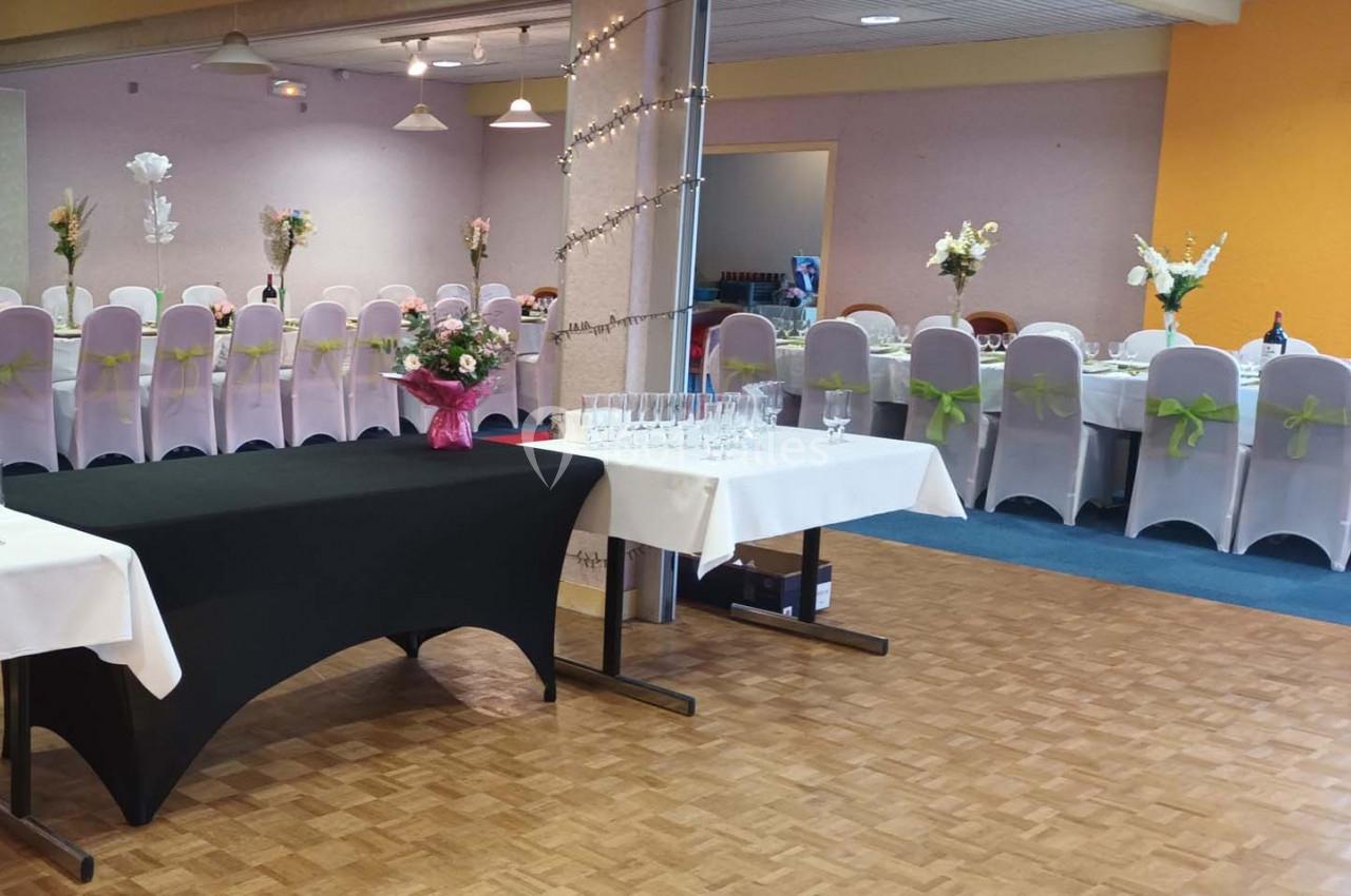 Salle décorée pour un événement avec tables dressées, chaises ornées de nœuds verts et fleurs en centre de table.