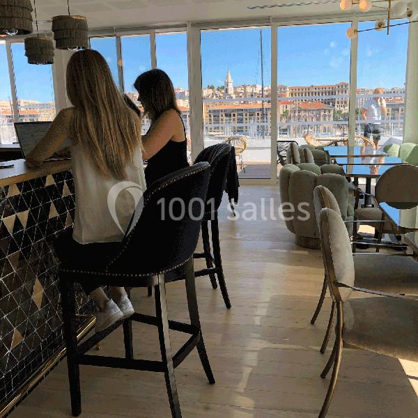 Deux femmes assises sur des tabourets dans un café lumineux avec vue sur une ville en arrière-plan.