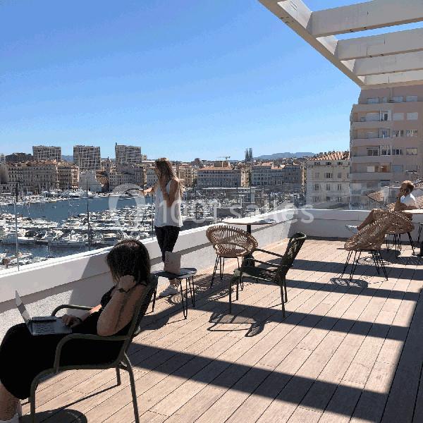 Personnes assises ou debout sur une terrasse ensoleillée avec vue sur un port et des immeubles en arrière-plan.