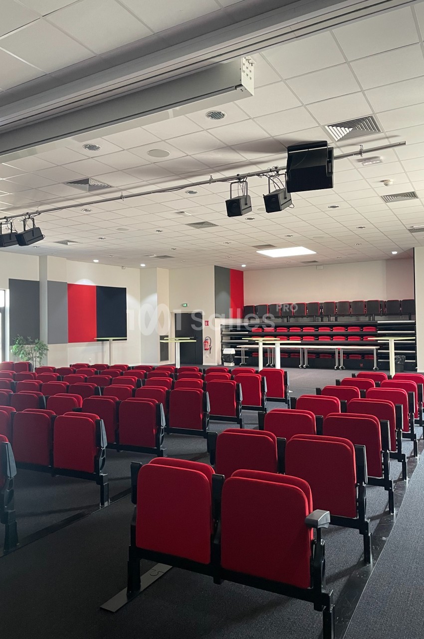 Salle de conférence vide avec des rangées de sièges rouges, une scène surélevée et un plafond équipé de projecteurs.