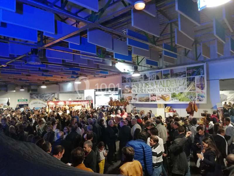 Foule rassemblée dans un espace intérieur éclairé, avec des stands et des panneaux promotionnels en arrière-plan.