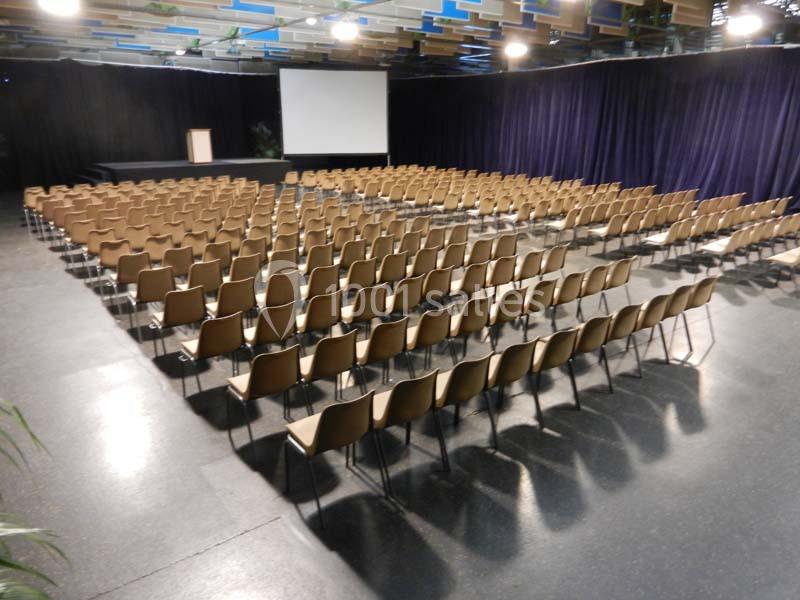 Salle de conférence avec des rangées de chaises en bois disposées face à un écran et un pupitre.