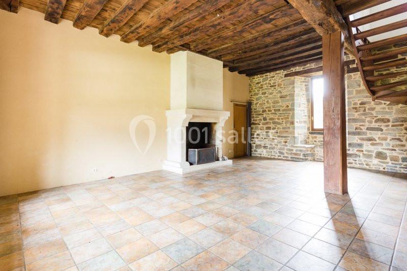 Location salle Domagné (Ille-et-Vilaine) - Domaine de Neuville #10 Salle vide avec cheminée en pierre, murs en pierre et plâtre, plafond en poutres apparentes et sol en carrelage.