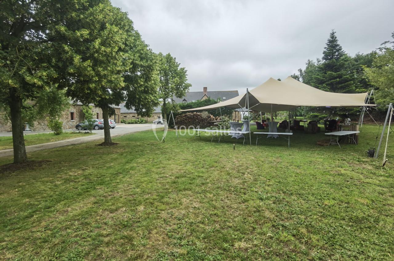 Location salle Domagné (Ille-et-Vilaine) - Domaine de Neuville #4 Tente beige installée sur une pelouse avec des tables dressées, entourée d'arbres et de bâtiments en arrière-plan.