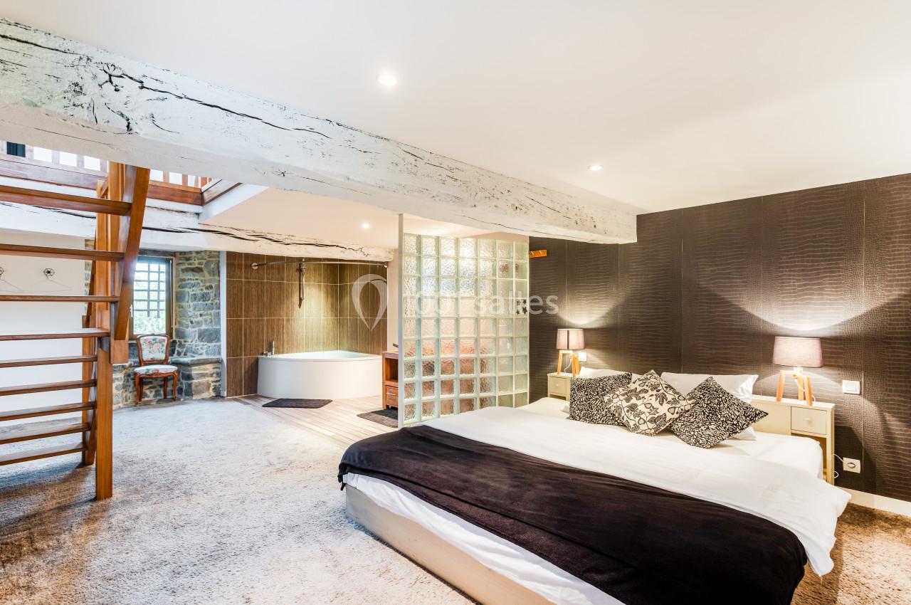 Location salle Domagné (Ille-et-Vilaine) - Domaine de Neuville #3 Chambre spacieuse avec lit double, poutres apparentes, baignoire en fond et escalier en bois menant à un étage supérieur.